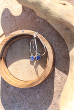 Load image into Gallery viewer, Silver hoop earrings with blue opal heart stones on a wooden stand, placed on sandy ground.