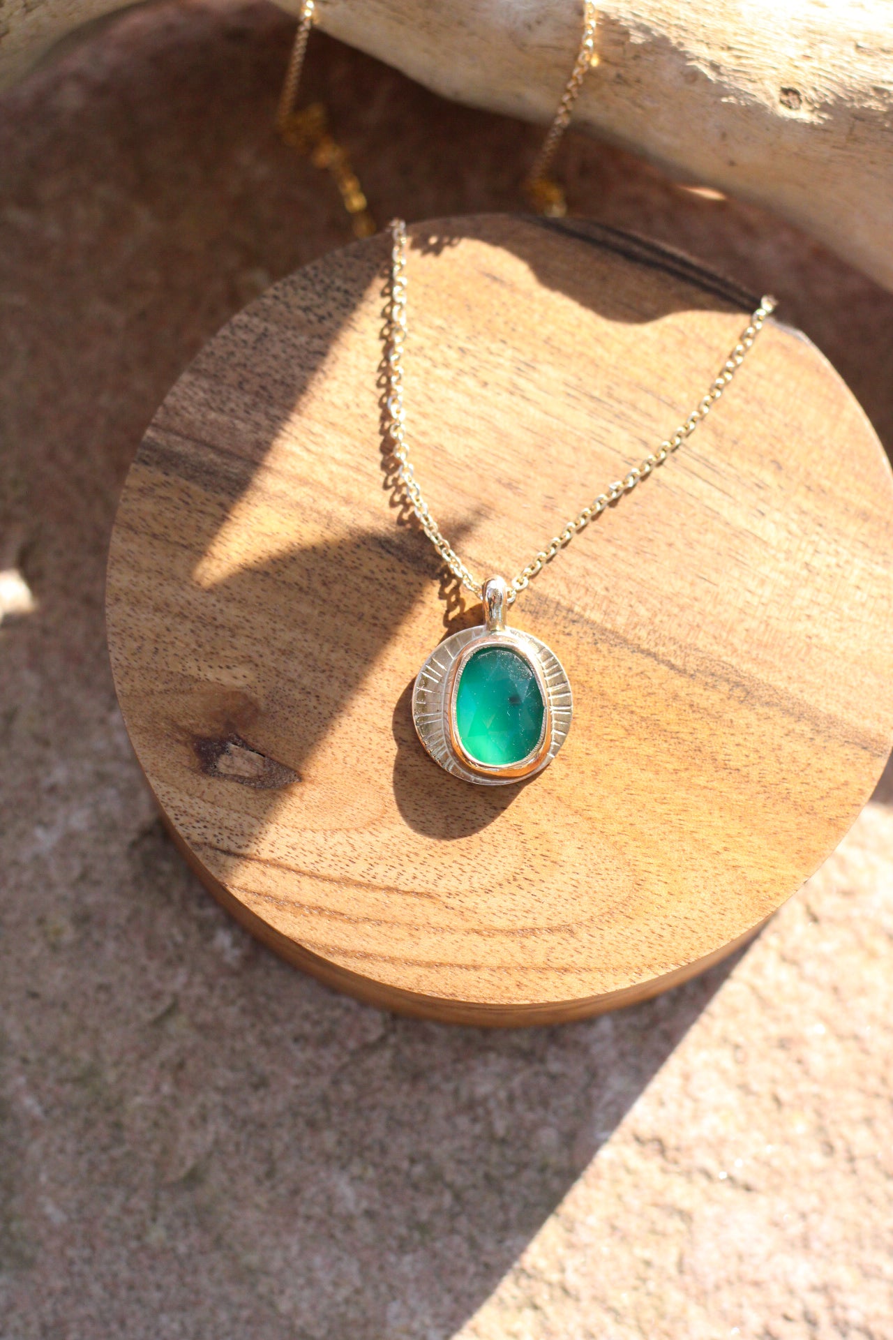 bright green stone against gold pendant with Iines, similar to sun rays. On a piece of wood upon a rock.