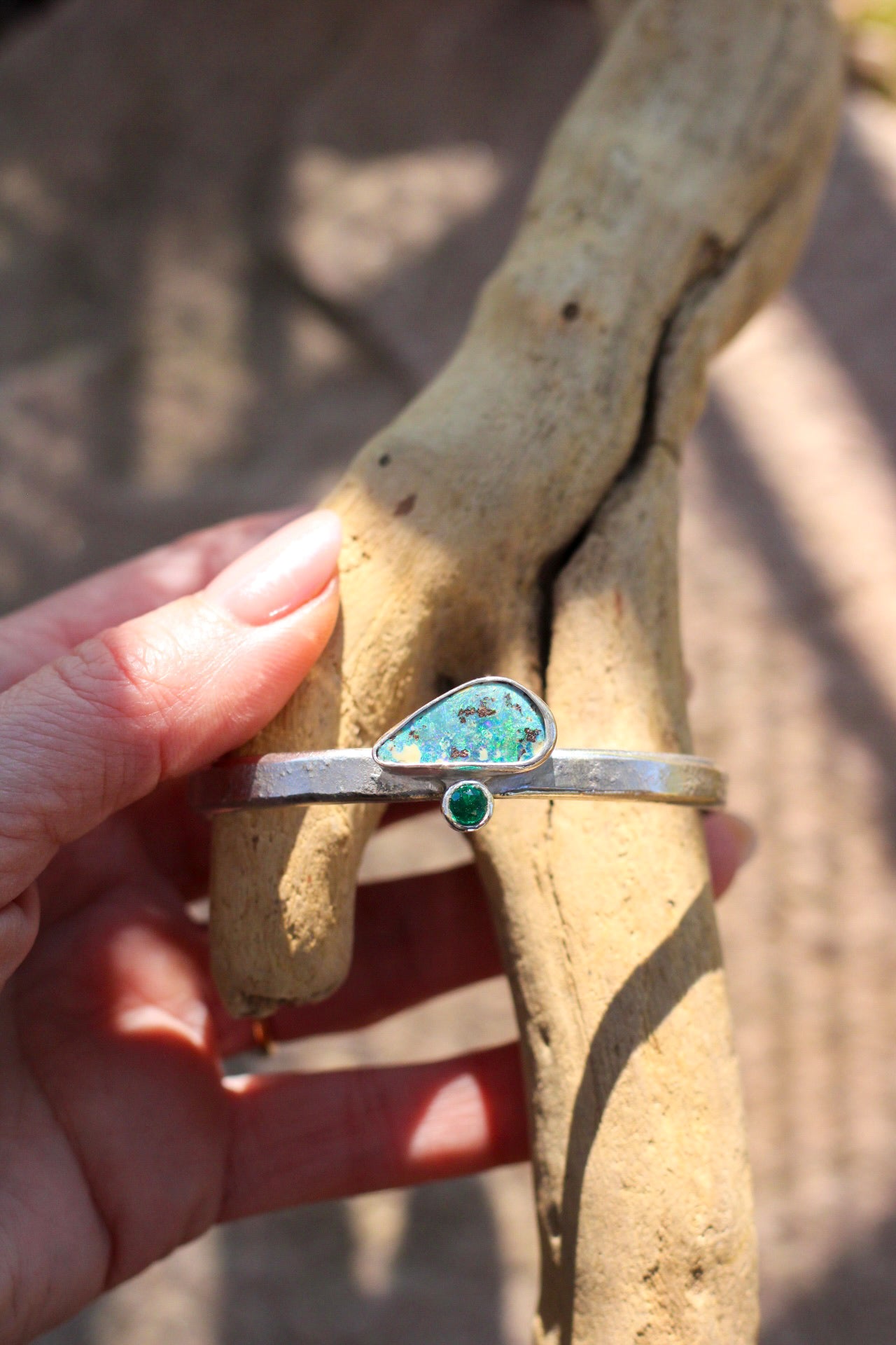 Silver bracelet with opal and emerald stone held against a tree branch