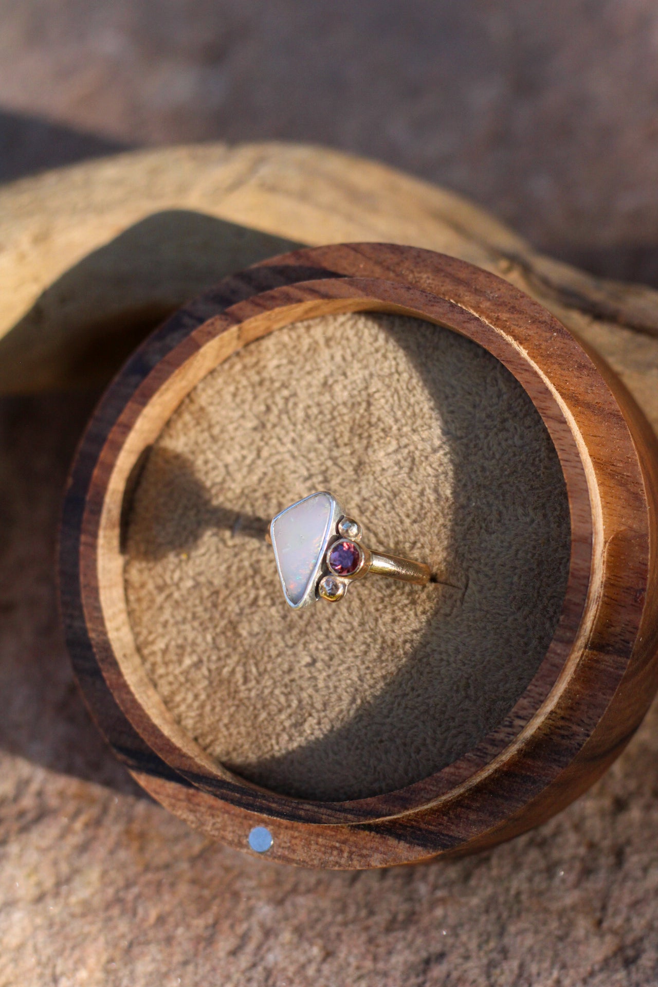 Ring with a triangular opal and round rose colored stone in a wooden box on a textured surface
