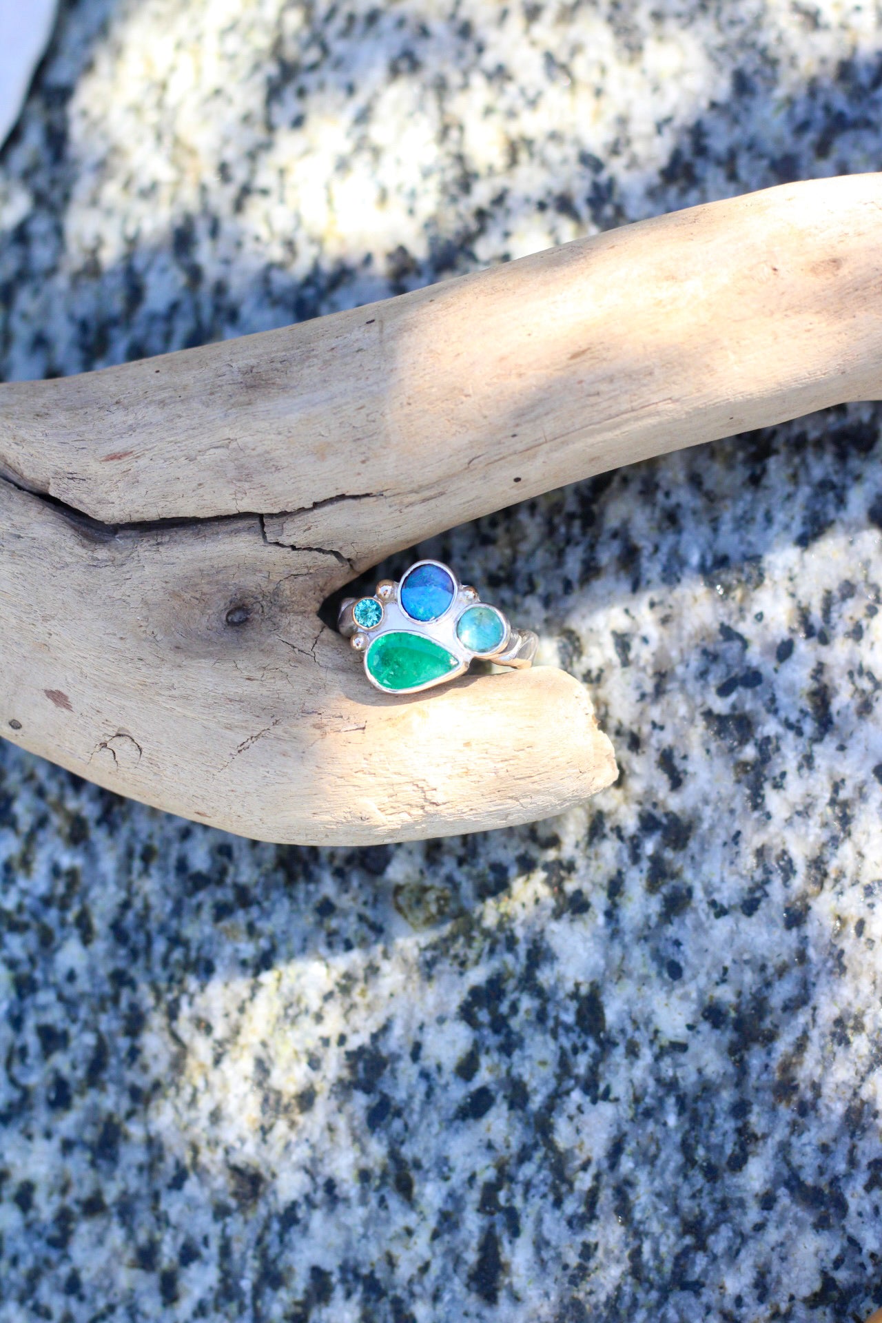 Ring with a collection of blue and green stones on a piece of driftwood against a rocky background